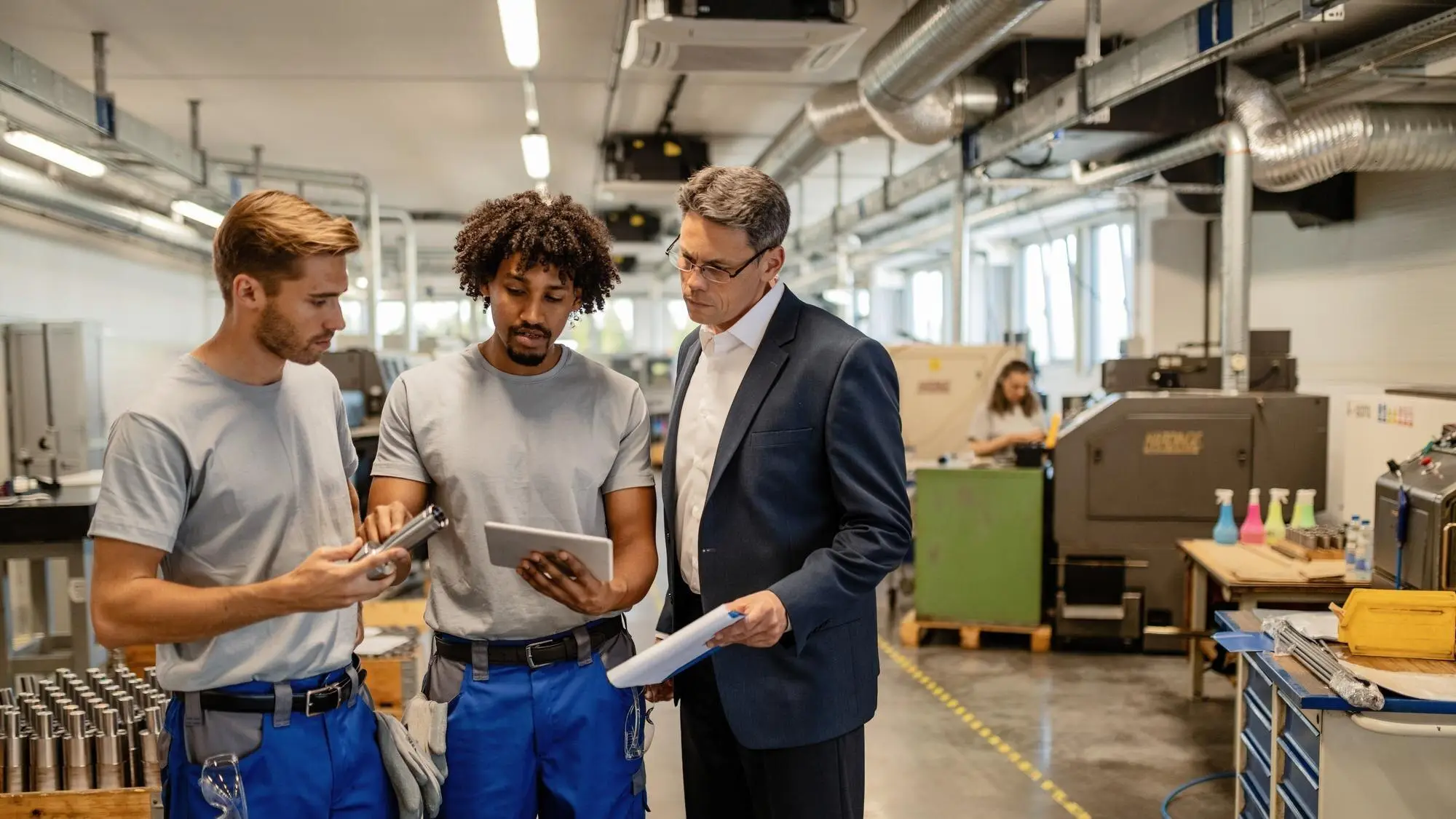 Ein Betriebsleiter und zwei Mitarbeiter besprechen in der Fertigungshalle Daten auf einem Tablet