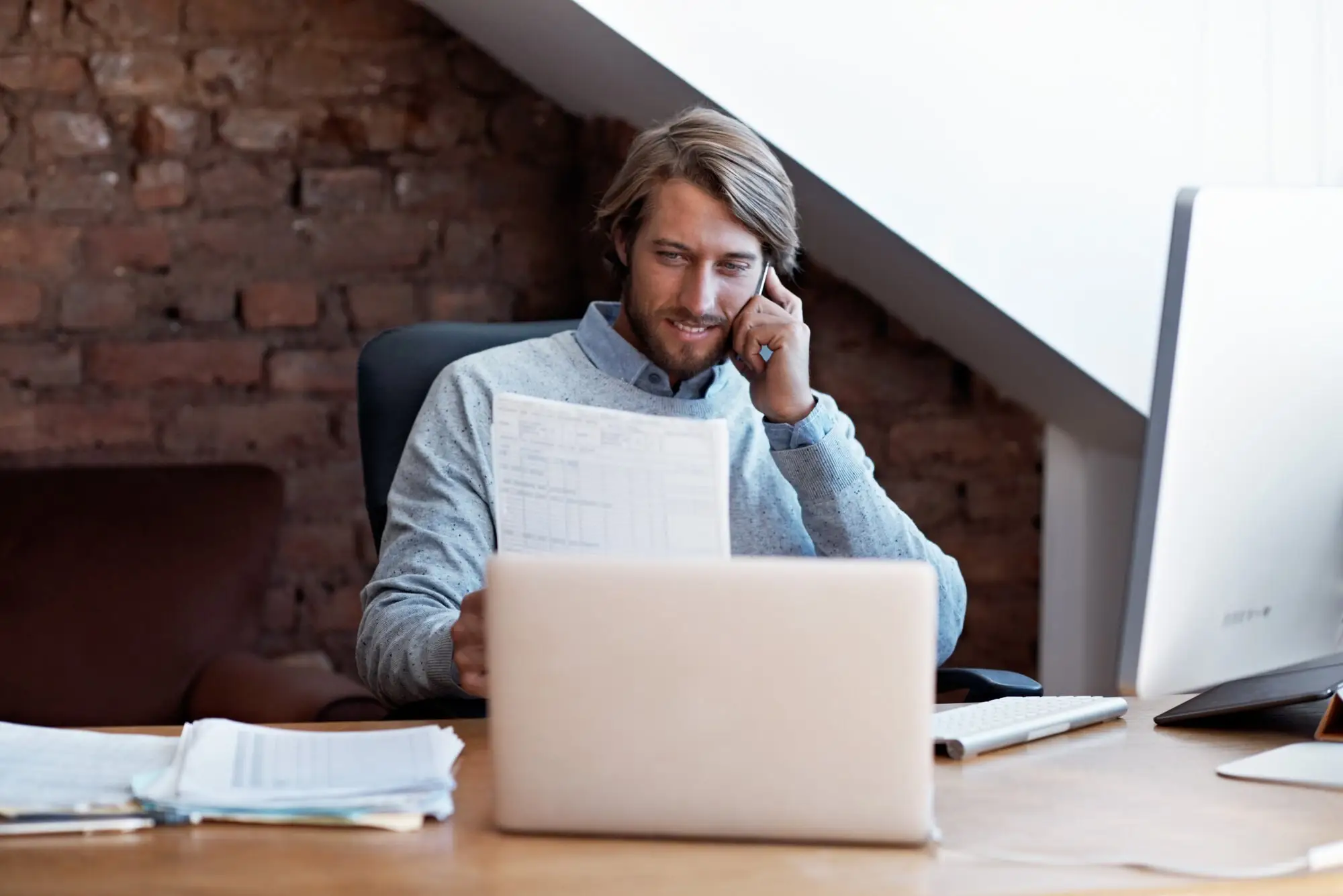 Mann telefoniert am Laptop und hält ein Blatt mit Notizen